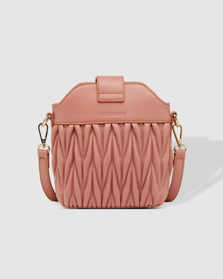 A woman outdoors holds the Louenhide Layla Crossbody Bag, a pink vegan leather mini bucket bag, paired with a one-shoulder striped top and white high-waisted pants.