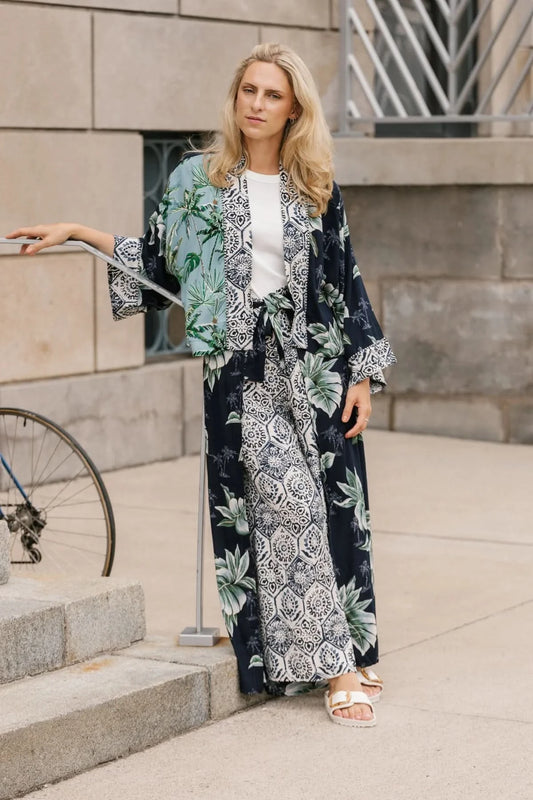 A woman stands on a sidewalk wearing the Shannon Passero Palm Cardi and white sandals, with part of a bicycle visible on the left.