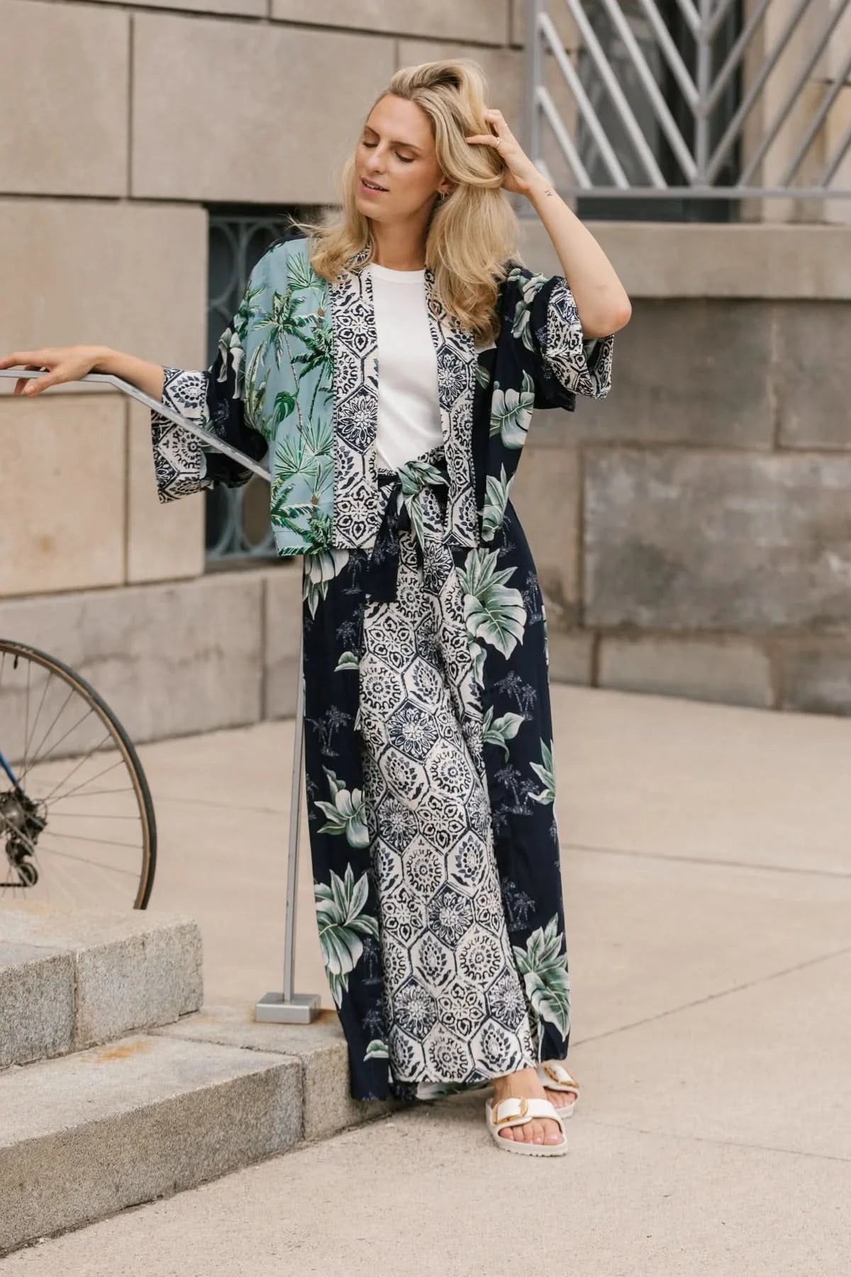 A woman stands on a sidewalk wearing the Shannon Passero Palm Cardi and white sandals, with part of a bicycle visible on the left.