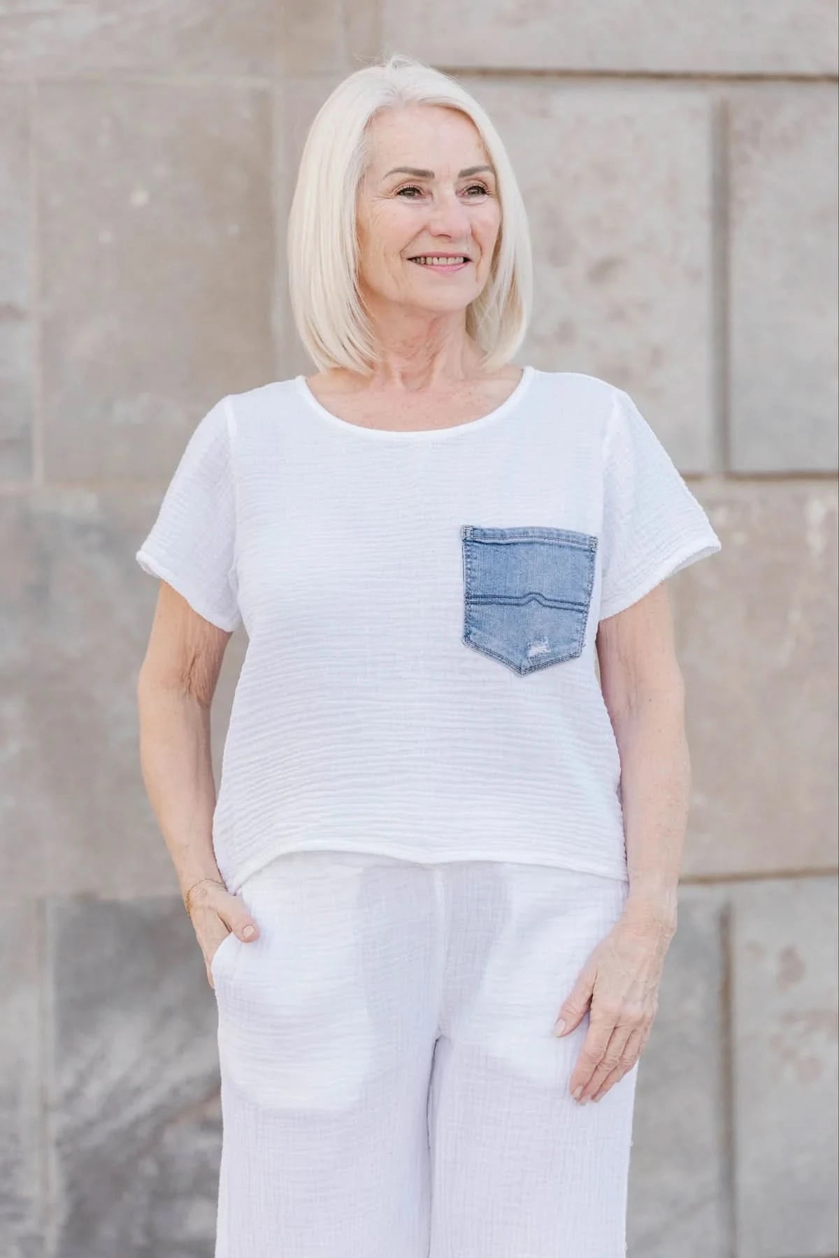 An older woman with straight white hair wears the Aletta Top by Shannon Passero, a cotton gauze shirt with a blue upcycled vintage denim pocket, paired with white pants and standing against a stone wall.