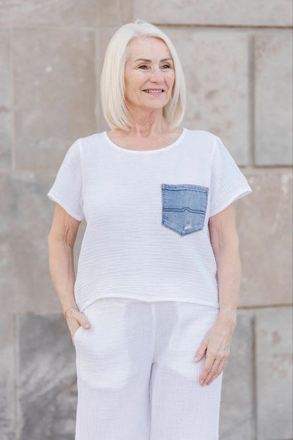 An older woman with straight white hair wears the Aletta Top by Shannon Passero, a cotton gauze shirt with a blue upcycled vintage denim pocket, paired with white pants and standing against a stone wall.