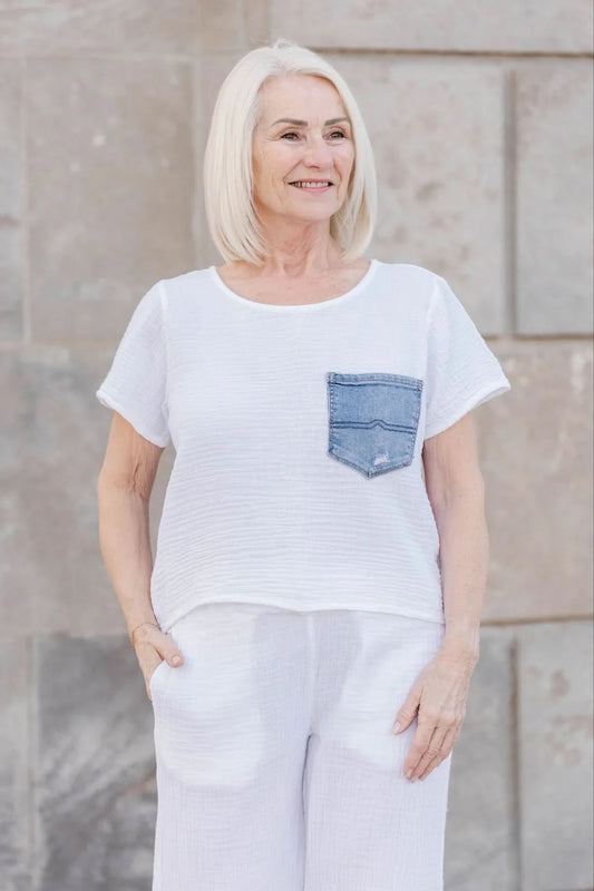 An older woman with straight white hair wears the Aletta Top by Shannon Passero, a cotton gauze shirt with a blue upcycled vintage denim pocket, paired with white pants and standing against a stone wall.