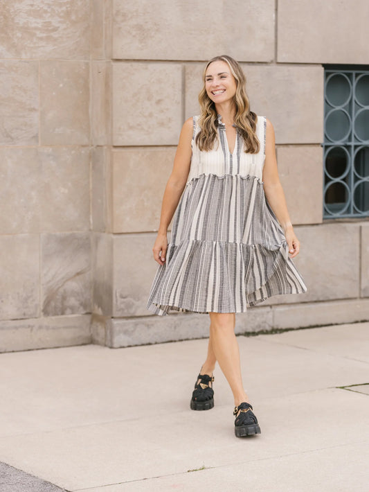 A person with long hair stands outdoors in a playful, v-neck, sleeveless Shannon Passero Whitby Tank Dress, smiling slightly.