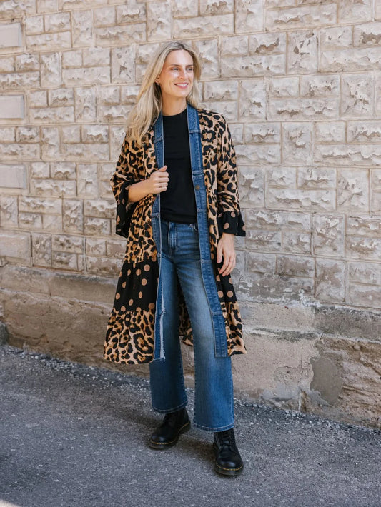 A woman stands against a stone wall wearing a black shirt, upcycled denim jeans, black boots, and the Shannon Passero Ying Cardigan—an eye-catching long coat with animal print and polka dots.
