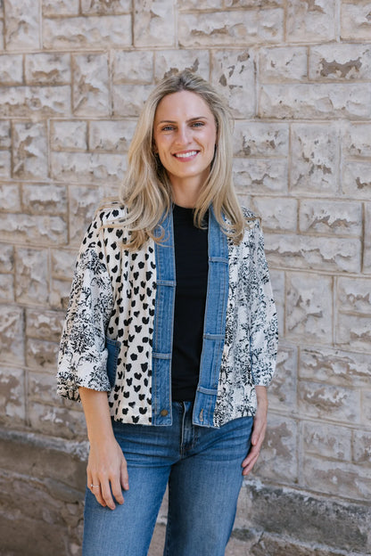 A woman with long blonde hair wears the Shannon Passero Karol Cardigan, a black shirt, and upcycled vintage denim jeans as she stands smiling in front of a textured stone wall, highlighting her sustainable style.