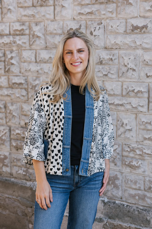 A woman with long blonde hair wears the Shannon Passero Karol Cardigan, a black shirt, and upcycled vintage denim jeans as she stands smiling in front of a textured stone wall, highlighting her sustainable style.