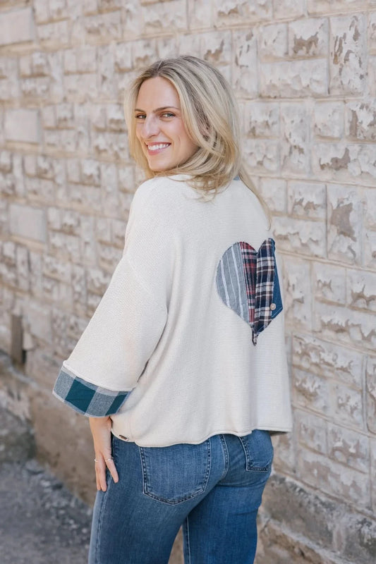 A woman stands sideways against a stone wall wearing the Alpine Love Cardigan by Shannon Passero—an upcycled flannel with a plaid heart patch on the back, plaid sleeve details, and blue jeans for a chic, sustainable look.