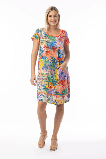 A woman stands smiling, wearing the Orientique Shift Dress—a colorful short-sleeve, knee-length organic cotton summer dress with pockets—paired with tan wedge sandals, set against a white background.