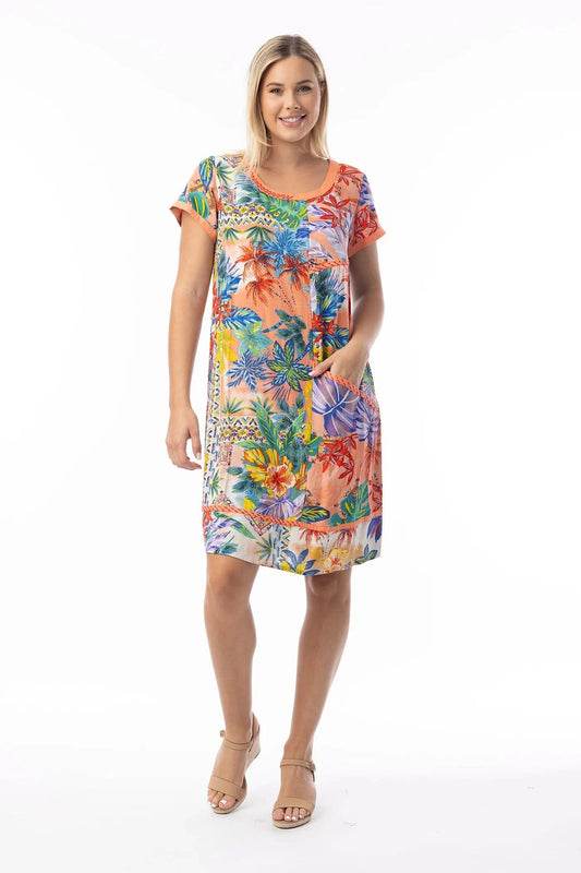 A woman stands smiling, wearing the Orientique Shift Dress—a colorful short-sleeve, knee-length organic cotton summer dress with pockets—paired with tan wedge sandals, set against a white background.
