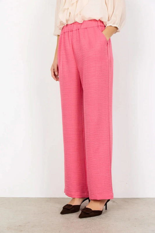 Model wearing Soya Concepts Charissa Pant in pink, styled with a light long-sleeve blouse and dark pointed-toe heels, standing against a plain white wall.