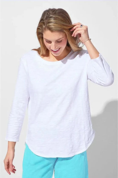 A woman wearing the Escape Peruvian Cotton Weekender Tee in white and light blue pants stands against a plain light background, looking down as she touches her hair.