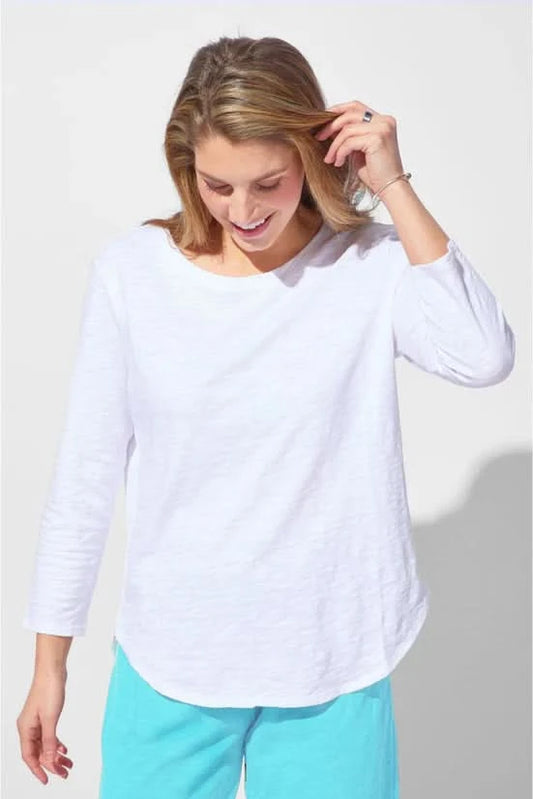 A woman wearing the Escape Peruvian Cotton Weekender Tee in white and light blue pants stands against a plain light background, looking down as she touches her hair.