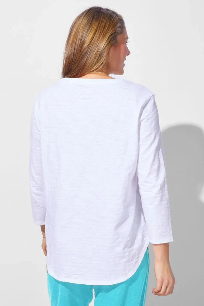 A woman wearing the Escape Peruvian Cotton Weekender Tee in white and light blue pants stands against a plain light background, looking down as she touches her hair.