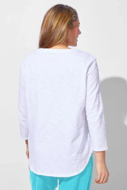 A woman wearing the Escape Peruvian Cotton Weekender Tee in white and light blue pants stands against a plain light background, looking down as she touches her hair.