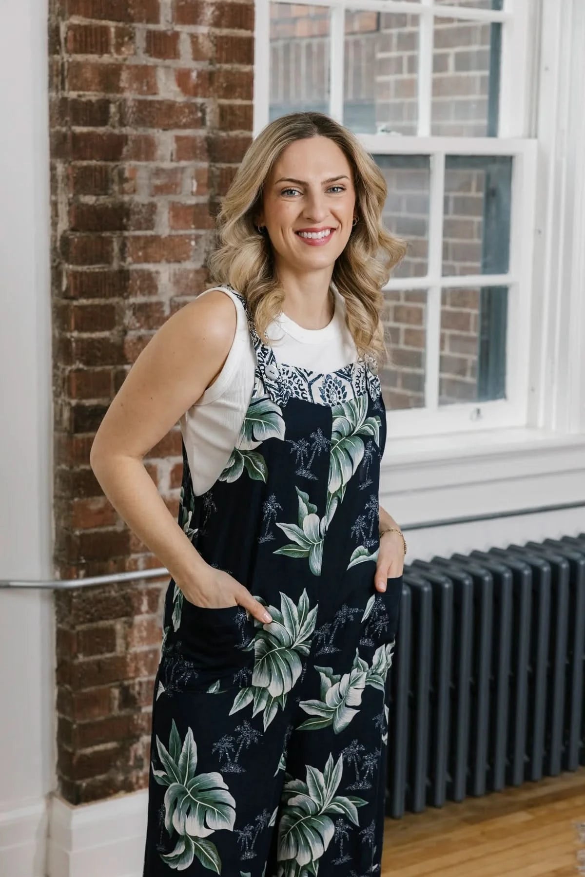 A woman with wavy blonde hair stands indoors by a brick wall, smiling with her hands in her pockets while wearing the Shannon Passero Elora Overall—floral, upcycled vintage denim—for a stylish and sustainable look.