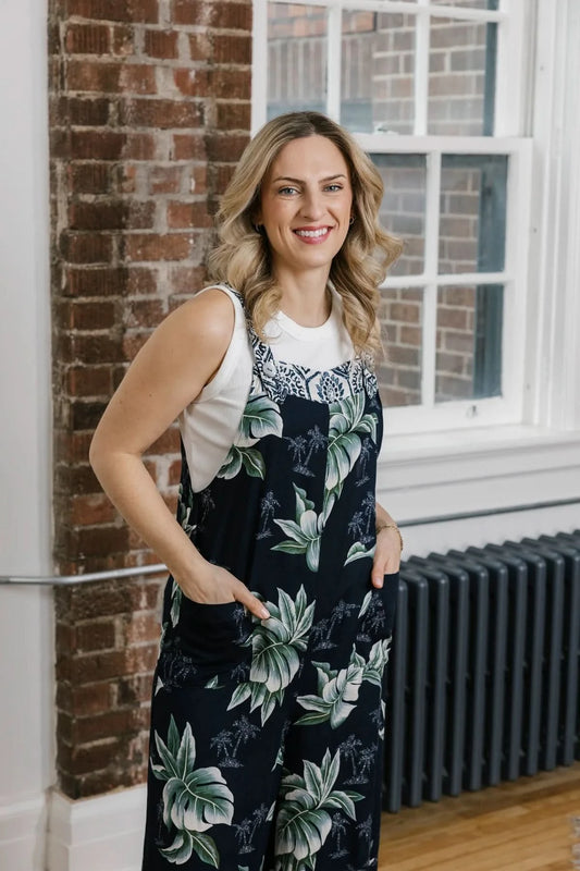 A woman with wavy blonde hair stands indoors by a brick wall, smiling with her hands in her pockets while wearing the Shannon Passero Elora Overall—floral, upcycled vintage denim—for a stylish and sustainable look.