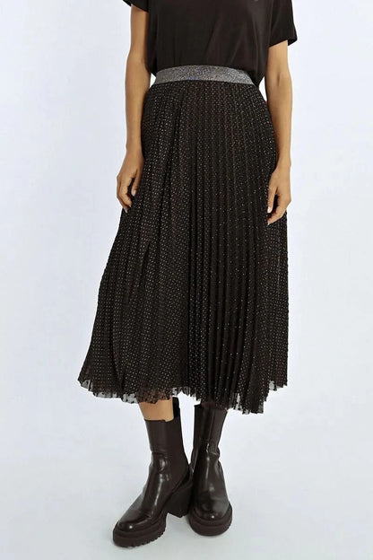 A person poses against a red backdrop, wearing a black top and the Pleated Plumetis Skirt by Molly Bracken, adorned with polka dot feathers, completed by silver heeled boots.