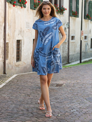 A woman in the Cherishh Beige Leafe Print Dress and sandals walks on a cobblestone street past an old building with green shutters and flower boxes.