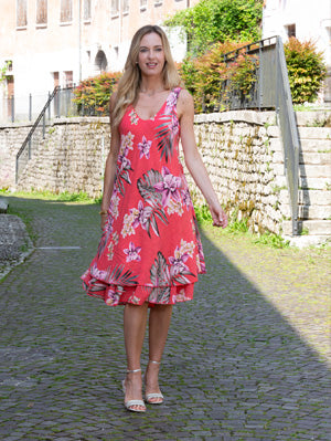 A woman wears the Cherishh Coral Printed Dress and sandals, standing on a cobblestone path bordered by stone walls and greenery.