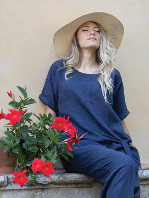A woman wearing the Cherishh Side Knot Top and a wide-brimmed hat sits by a beige wall near a red flowering plant, eyes closed and face tilted upward.