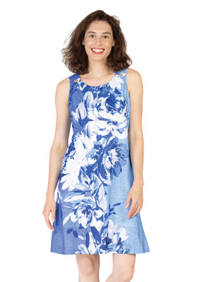 A woman stands smiling against a white background, wearing the Variations Rose Print Dress—a sleeveless, knee-length blue and white floral dress.