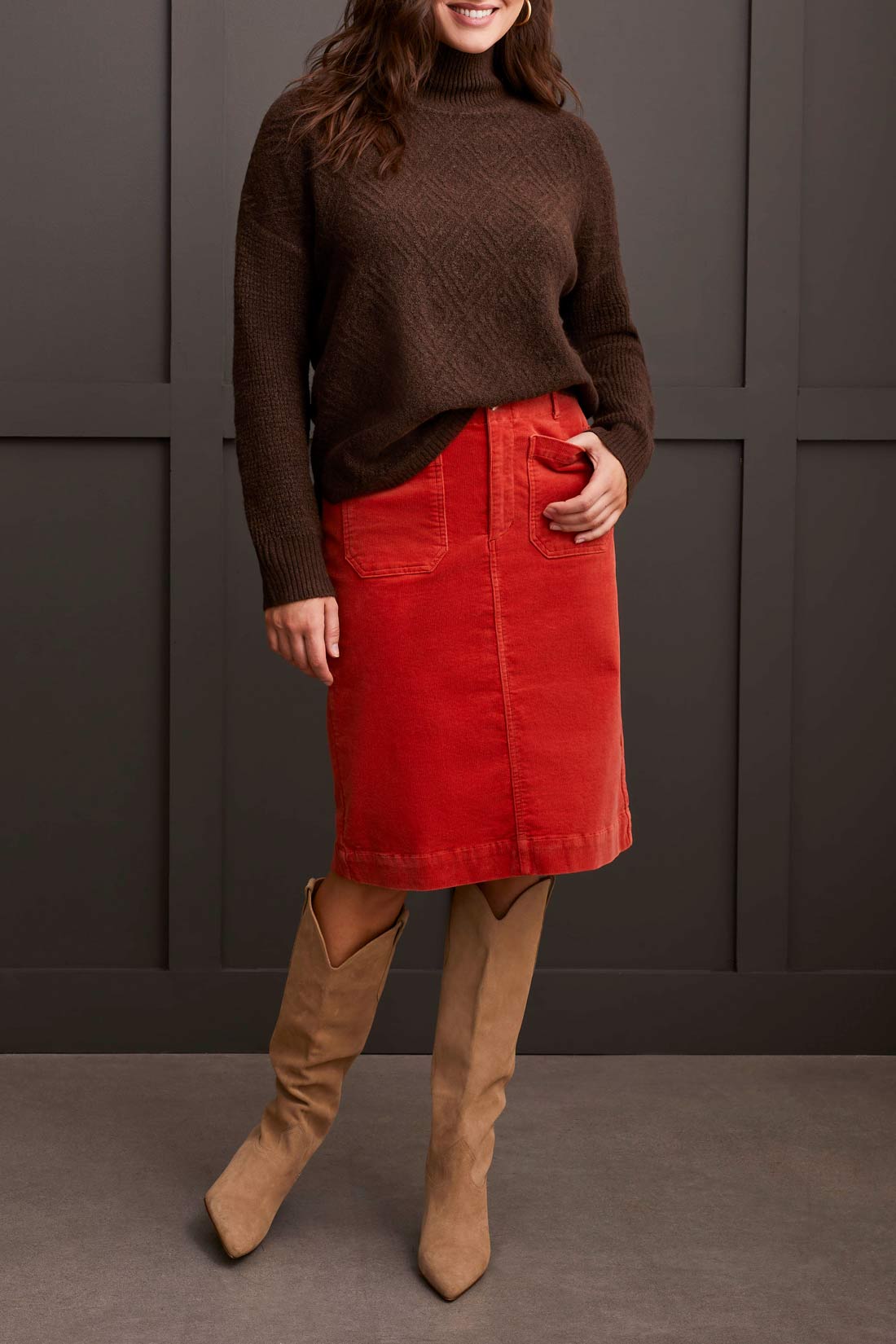 A person dressed in a cozy comfort ensemble featuring Tribal's coffee-colored Mock Neck Sweater, a red skirt, and tan knee-high boots stands against a dark paneled wall.