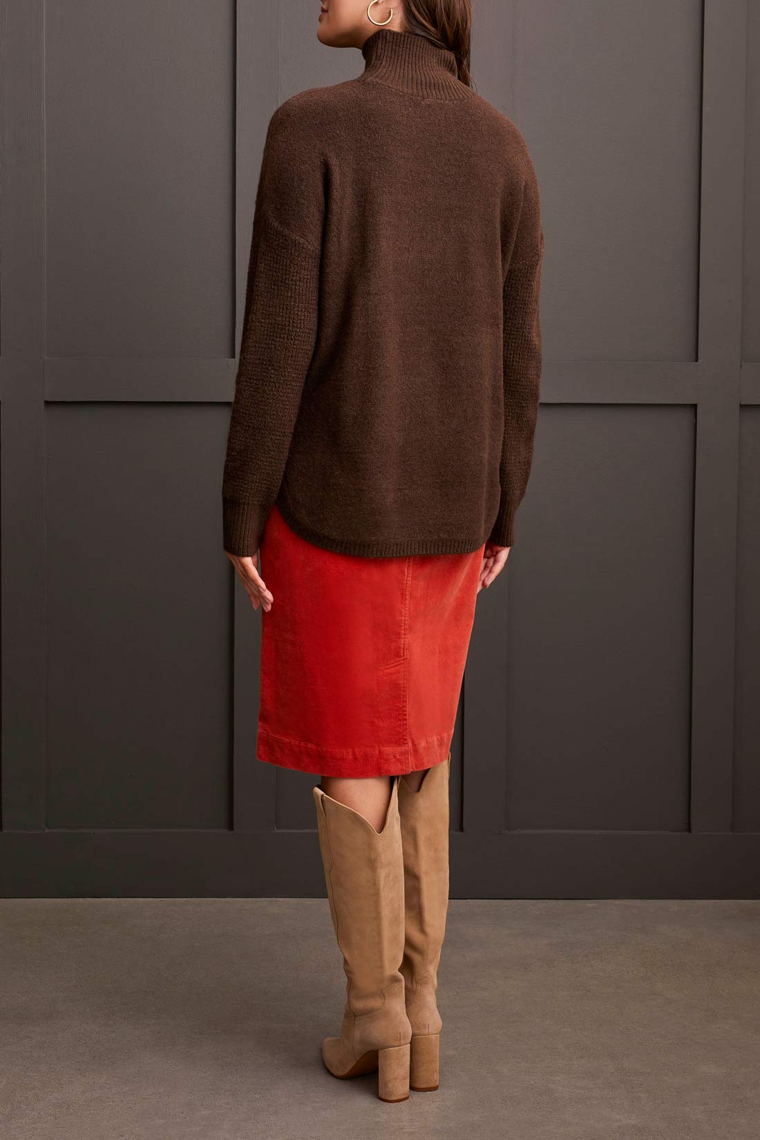 A person dressed in a cozy comfort ensemble featuring Tribal's coffee-colored Mock Neck Sweater, a red skirt, and tan knee-high boots stands against a dark paneled wall.