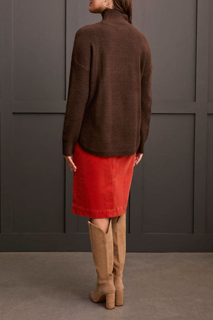 A person dressed in a cozy comfort ensemble featuring Tribal's coffee-colored Mock Neck Sweater, a red skirt, and tan knee-high boots stands against a dark paneled wall.