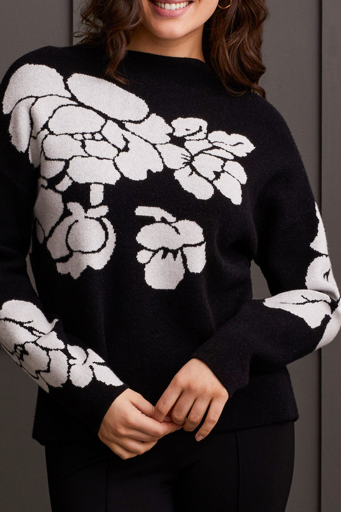 An individual is pictured from the shoulders down, donning a black Floral Funnel Neck Sweater by Tribal, adorned with a striking large white floral pattern. This cute fall sweater, made from cozy material, is ideal for cool autumn days. The person stands against a dark backdrop.