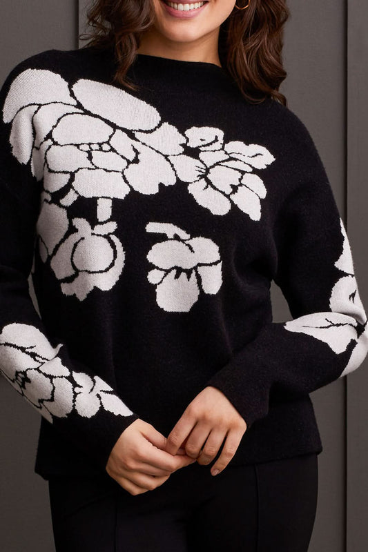 An individual is pictured from the shoulders down, donning a black Floral Funnel Neck Sweater by Tribal, adorned with a striking large white floral pattern. This cute fall sweater, made from cozy material, is ideal for cool autumn days. The person stands against a dark backdrop.