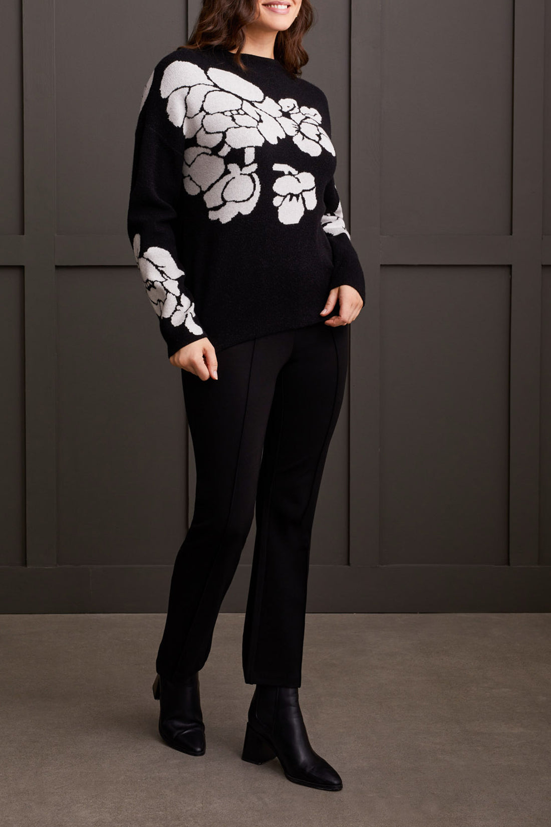 An individual is pictured from the shoulders down, donning a black Floral Funnel Neck Sweater by Tribal, adorned with a striking large white floral pattern. This cute fall sweater, made from cozy material, is ideal for cool autumn days. The person stands against a dark backdrop.