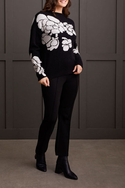 An individual is pictured from the shoulders down, donning a black Floral Funnel Neck Sweater by Tribal, adorned with a striking large white floral pattern. This cute fall sweater, made from cozy material, is ideal for cool autumn days. The person stands against a dark backdrop.