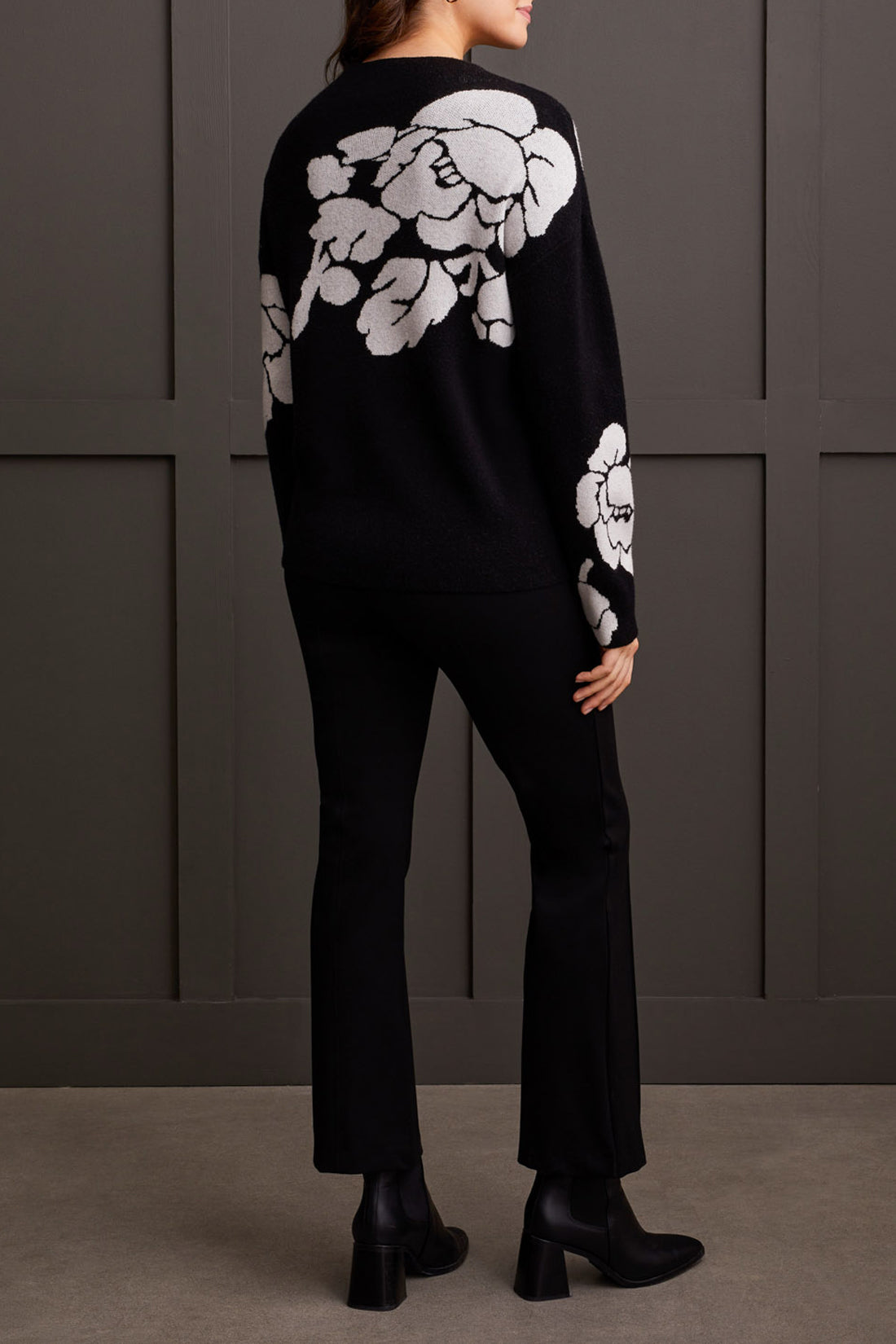 An individual is pictured from the shoulders down, donning a black Floral Funnel Neck Sweater by Tribal, adorned with a striking large white floral pattern. This cute fall sweater, made from cozy material, is ideal for cool autumn days. The person stands against a dark backdrop.