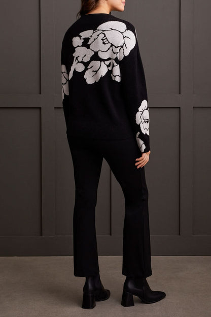 An individual is pictured from the shoulders down, donning a black Floral Funnel Neck Sweater by Tribal, adorned with a striking large white floral pattern. This cute fall sweater, made from cozy material, is ideal for cool autumn days. The person stands against a dark backdrop.
