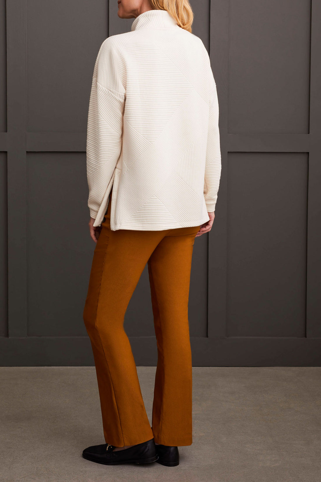 A person is wearing the Tribal Long Sleeve Tunic with Side Slits in white and orange-brown pants, standing against a dark gray paneled wall. This tunic, perfect for any fall wardrobe, showcases subtle side slits for added style. The person's head is cropped out of the image.