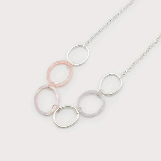The "Resin & Metal Rings on Chain" necklace by Caracol features interlocking rings in silver, pink, and purple hues, all set on a delicate chain against a white background.