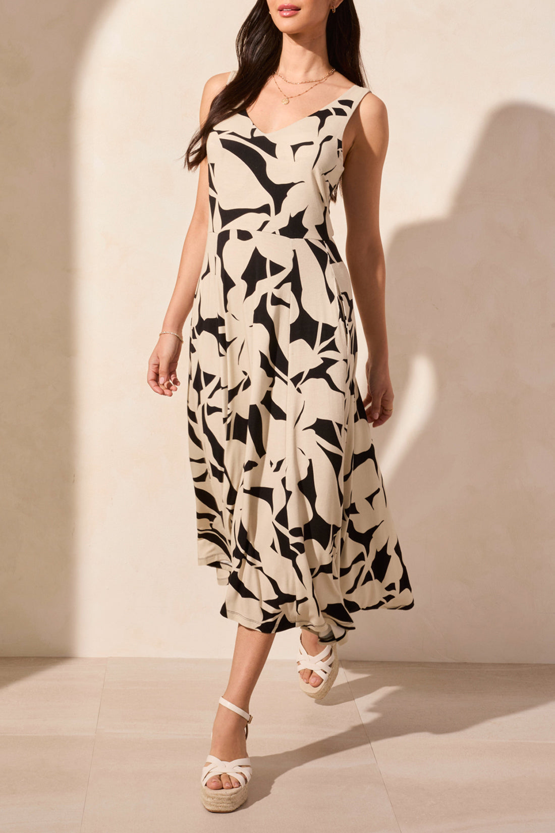 A woman stands against a light background, wearing Tribal's sleeveless high low dress featuring an abstract black and white print, paired with wedge sandals.