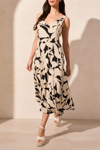 A woman stands against a light background, wearing Tribal's sleeveless high low dress featuring an abstract black and white print, paired with wedge sandals.