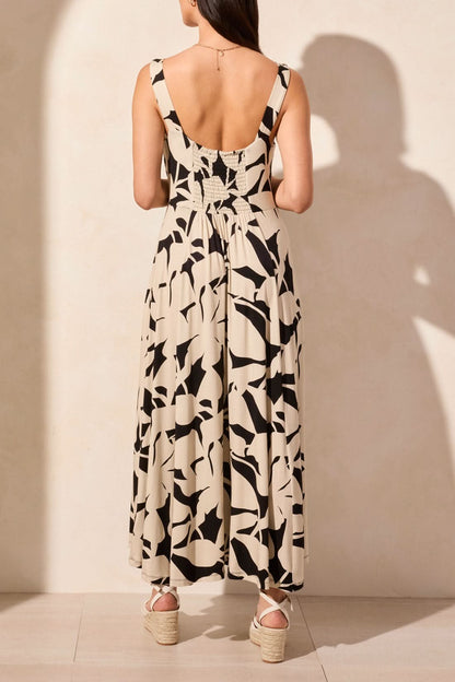 A woman stands against a light background, wearing Tribal's sleeveless high low dress featuring an abstract black and white print, paired with wedge sandals.