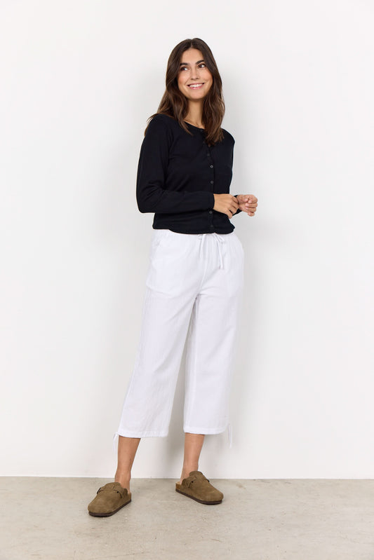 A woman stands against a white wall, smiling with her hands together. She wears a black long-sleeve shirt, brown slip-on shoes, and the Soya Concepts Cissie 1-C Cotton Pant—white cropped pants with an elastic waist for comfort.