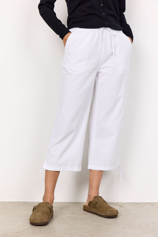A woman stands against a white wall, smiling with her hands together. She wears a black long-sleeve shirt, brown slip-on shoes, and the Soya Concepts Cissie 1-C Cotton Pant—white cropped pants with an elastic waist for comfort.