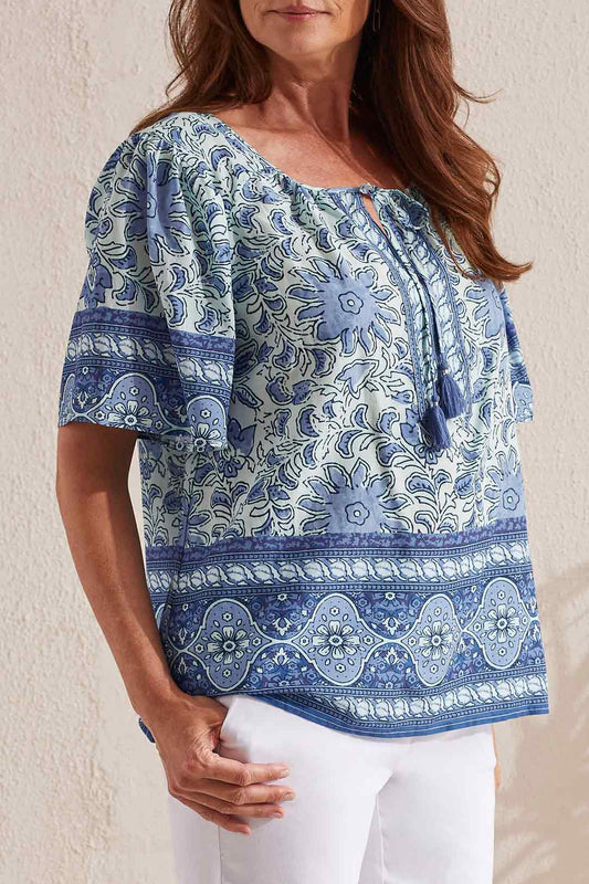 A woman wearing a Tribal blue and white patterned Short Sleeve Blouse with Tassel details standing against a neutral backdrop.