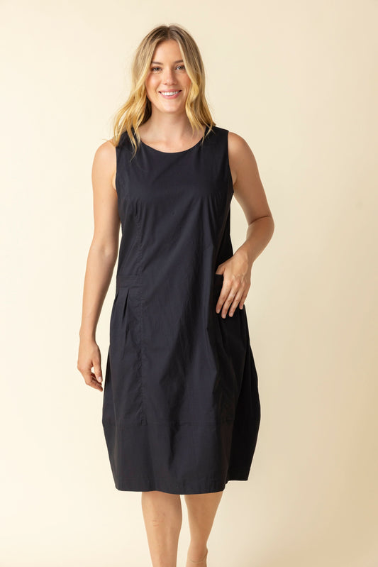 A woman smiles against a plain light background, wearing the Habitat Sleeveless Bubble Dress—a flattering silhouette—with one hand by her side and the other touching her dress.