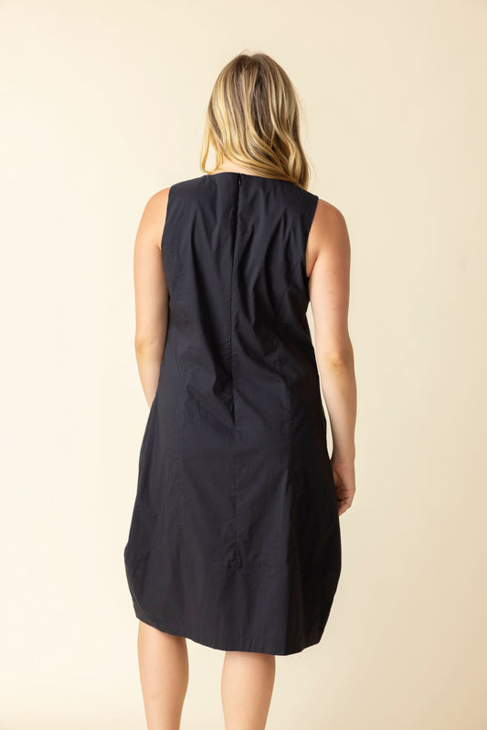 A woman smiles against a plain light background, wearing the Habitat Sleeveless Bubble Dress—a flattering silhouette—with one hand by her side and the other touching her dress.