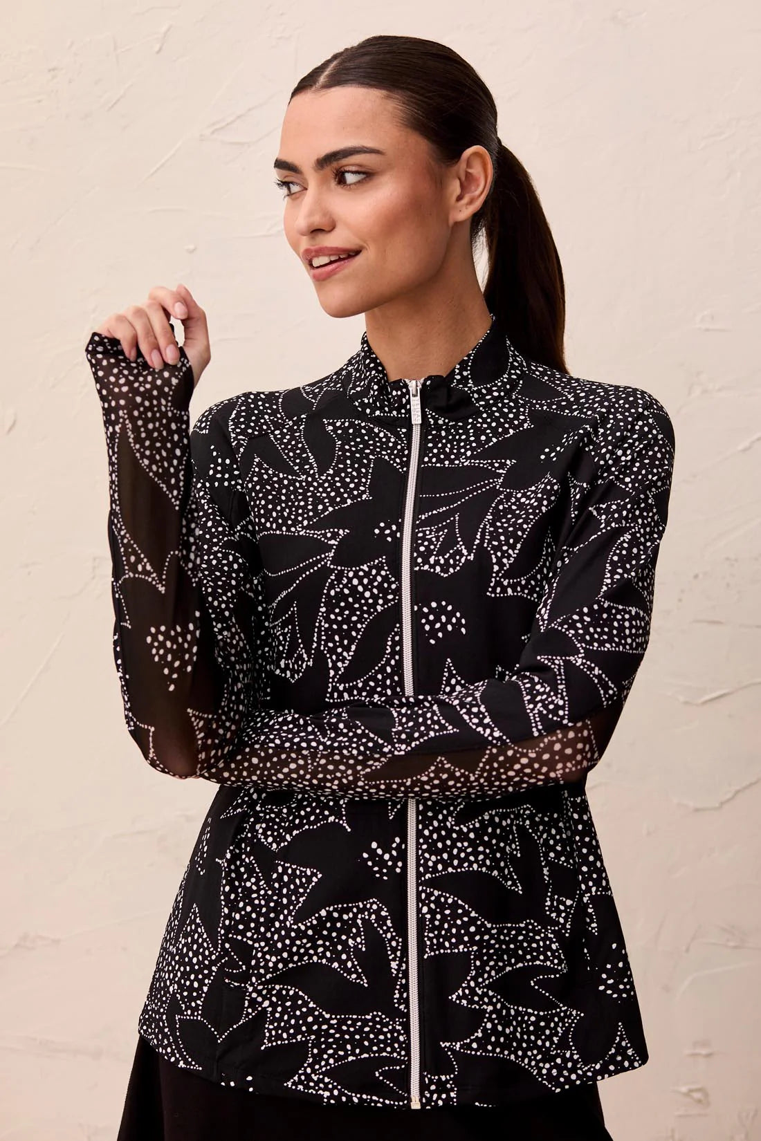 A woman wears the Tribal HIGH PERFORMANCE MESH MOCK-NECK ZIP-UP dress with a blue, green, and white leaf pattern and UPF 50+ protection, standing indoors with her hand in a pocket.