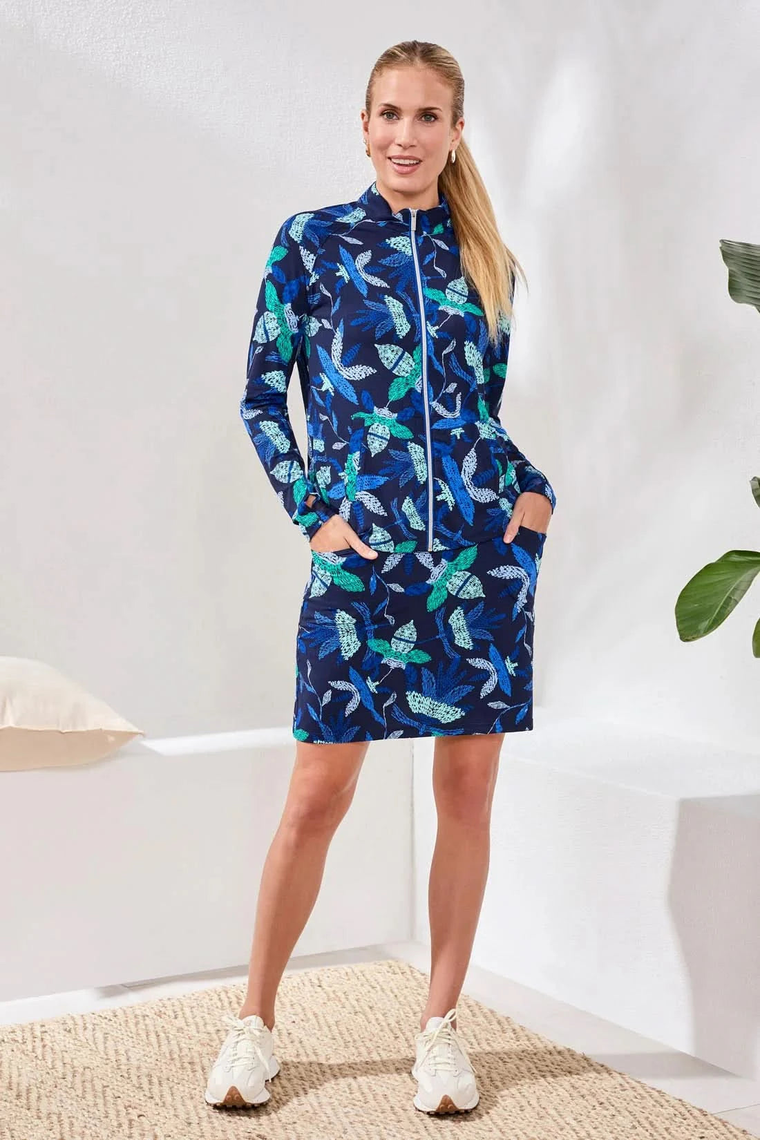A woman wears the Tribal HIGH PERFORMANCE MESH MOCK-NECK ZIP-UP dress with a blue, green, and white leaf pattern and UPF 50+ protection, standing indoors with her hand in a pocket.