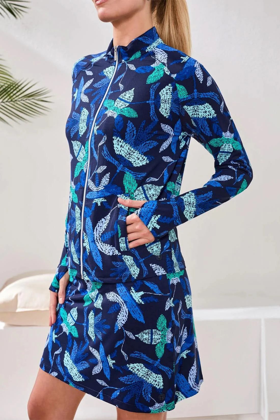 A woman wears the Tribal HIGH PERFORMANCE MESH MOCK-NECK ZIP-UP dress with a blue, green, and white leaf pattern and UPF 50+ protection, standing indoors with her hand in a pocket.