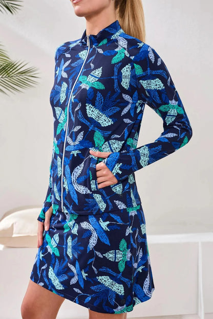A woman wears the Tribal HIGH PERFORMANCE MESH MOCK-NECK ZIP-UP dress with a blue, green, and white leaf pattern and UPF 50+ protection, standing indoors with her hand in a pocket.