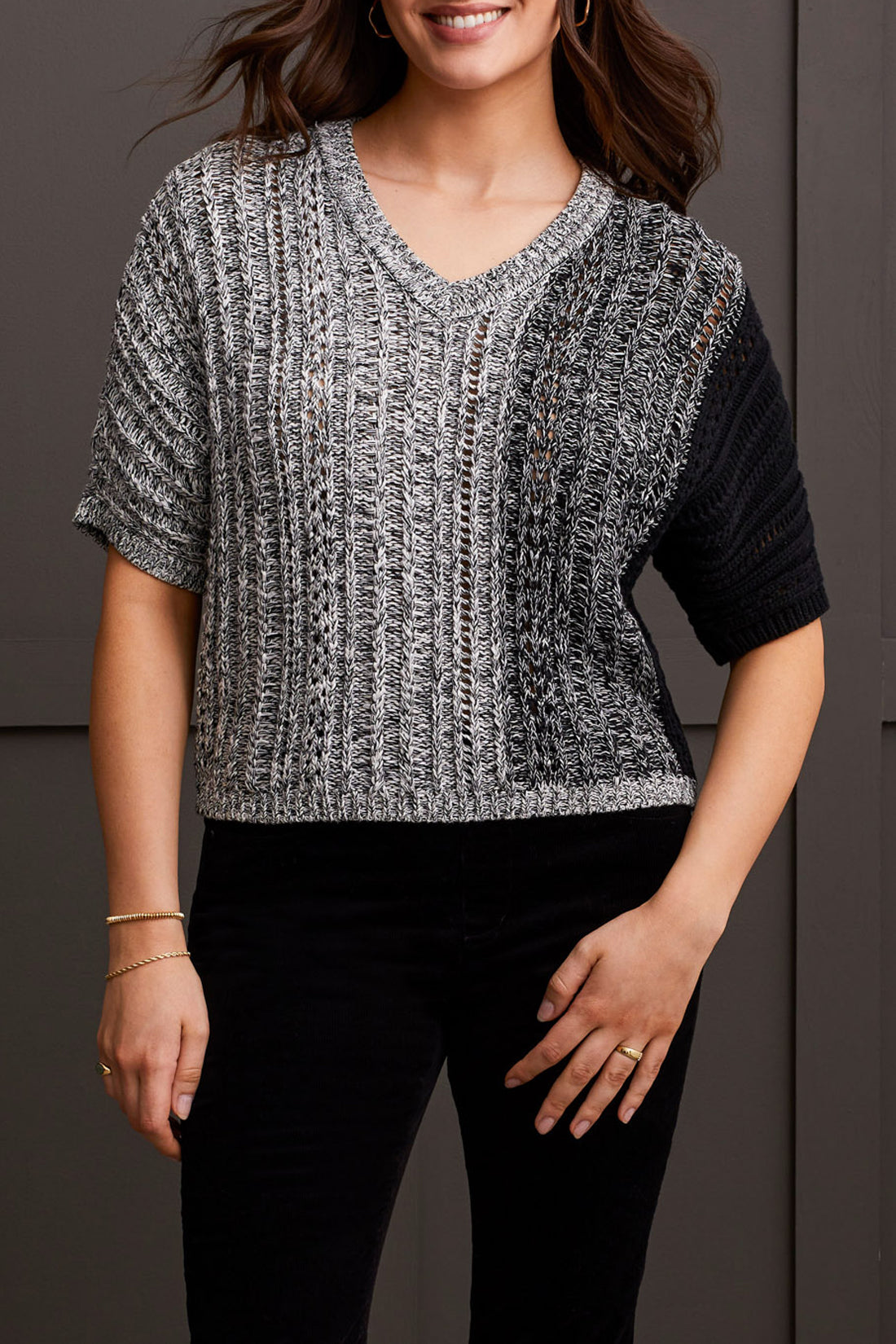 A person wearing the Tribal Dolman Elbow Sleeve Sweater, a cozy dual-toned knit featuring black and gray sections, short Dolman elbow sleeves, and a V-neckline, is paired with black pants and stands against a dark background.