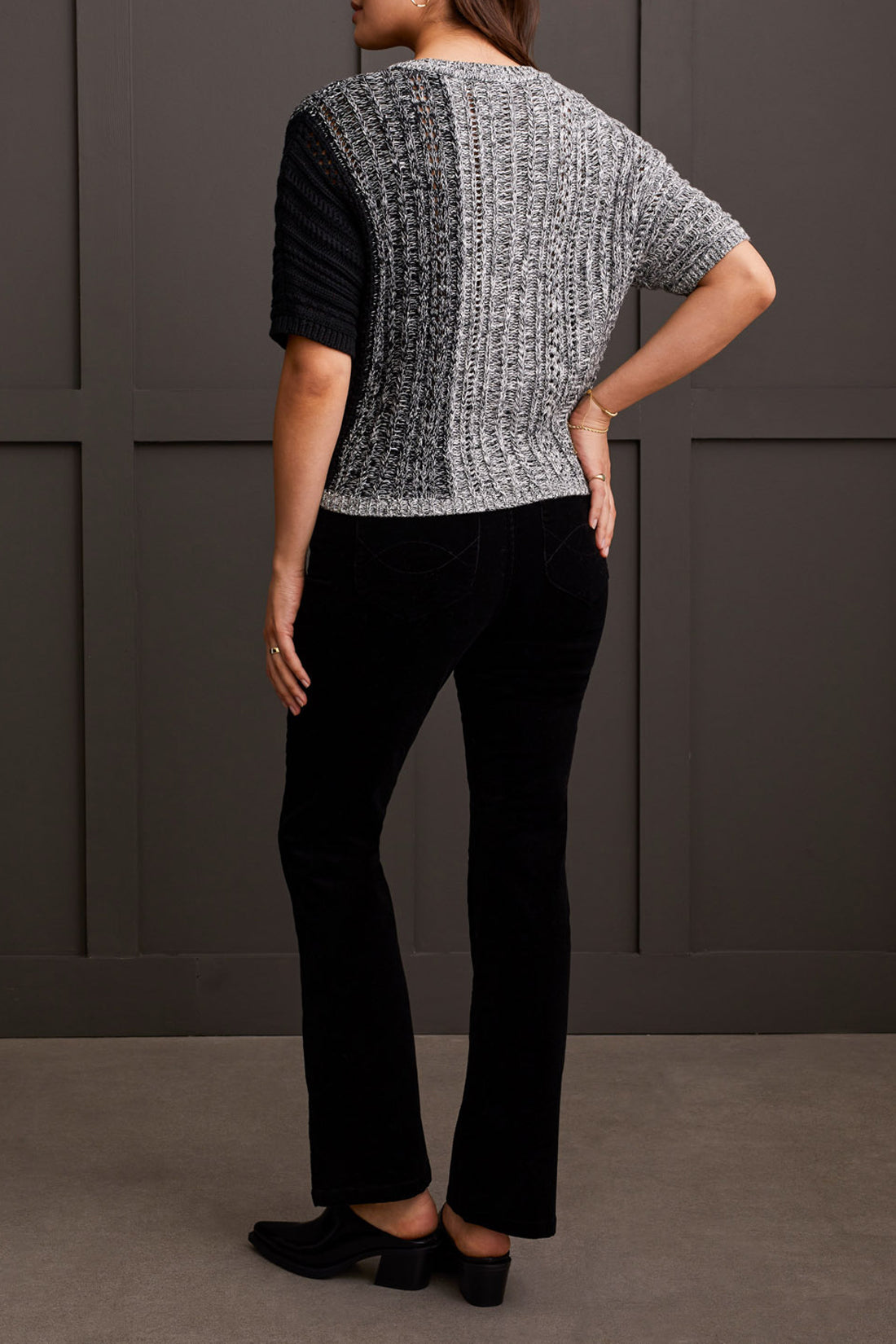 A person wearing the Tribal Dolman Elbow Sleeve Sweater, a cozy dual-toned knit featuring black and gray sections, short Dolman elbow sleeves, and a V-neckline, is paired with black pants and stands against a dark background.
